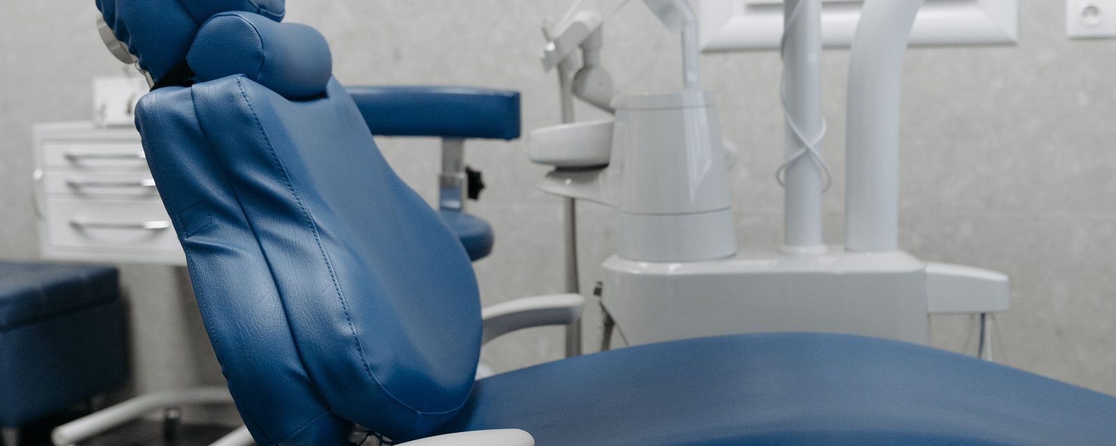 An empty blue dental chair in a treatment room, with equipment and a cabinet behind.