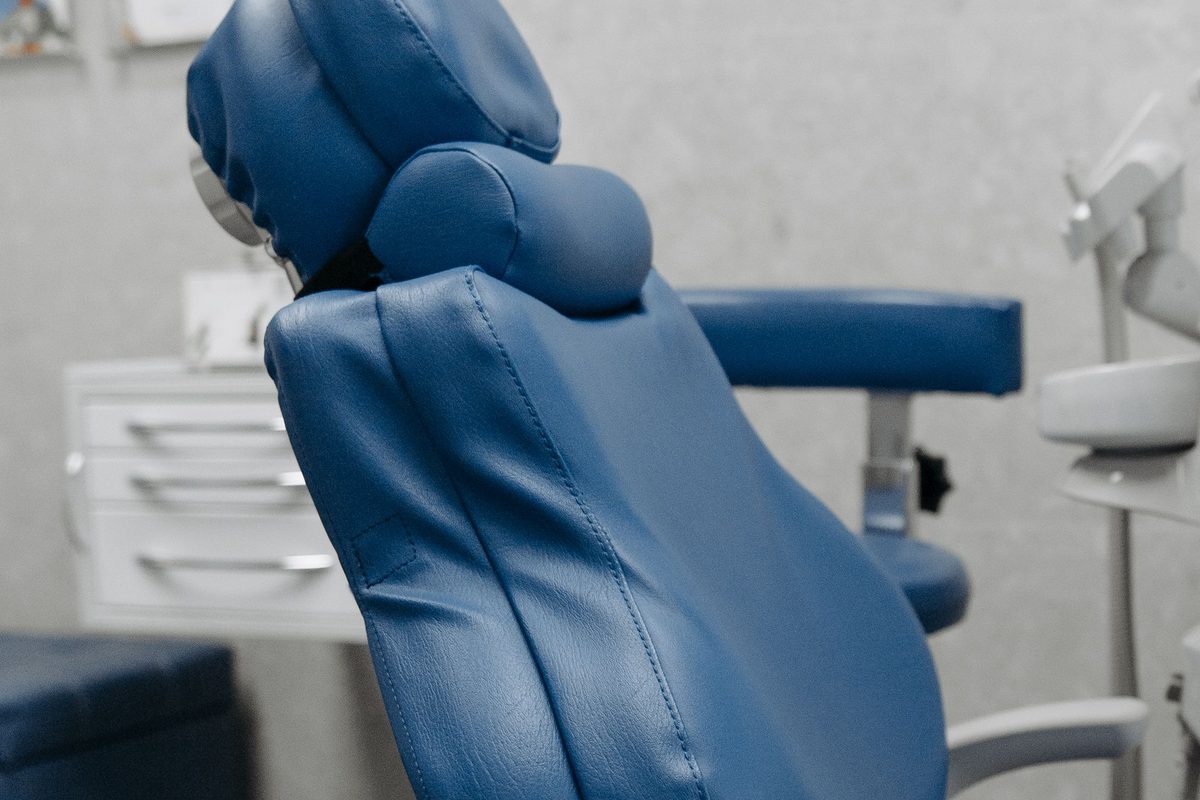 An empty blue dental chair in a treatment room, with equipment and a cabinet behind.