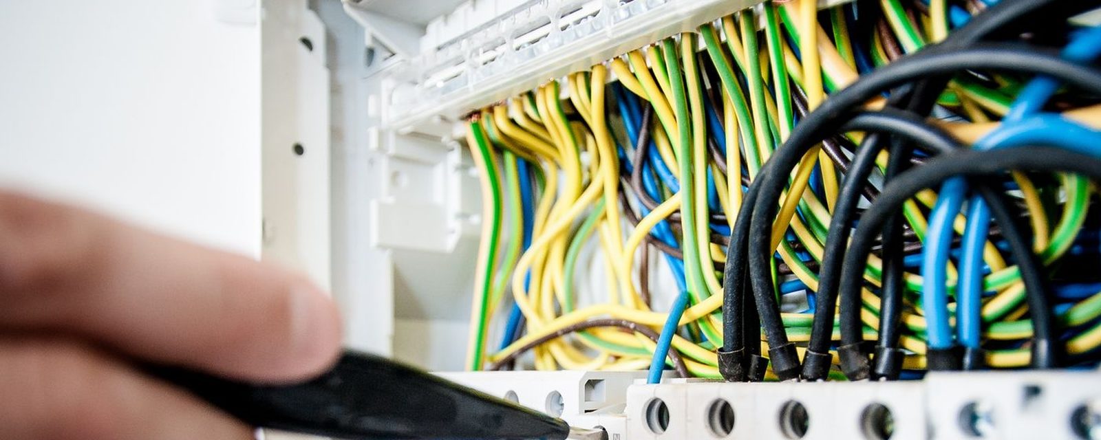 A hand with cable strippers working at an open panel of colour-coded electrical wiring.