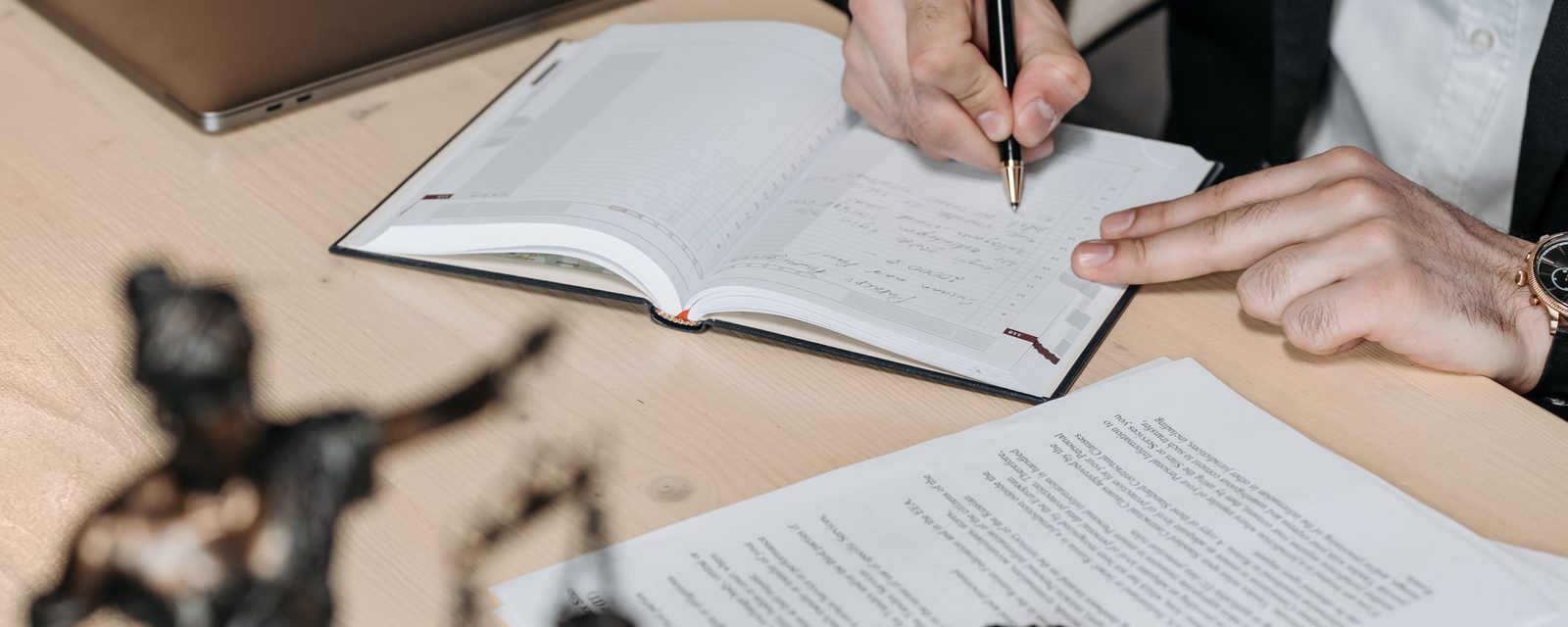 A hand writing in a diary at a desk, with papers and a pen.