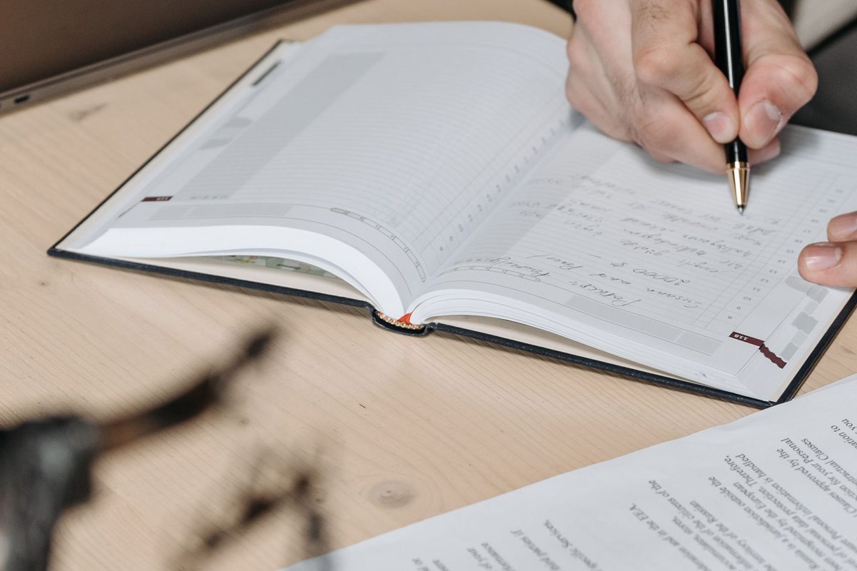 A hand writing in a diary at a desk, with papers and a pen.